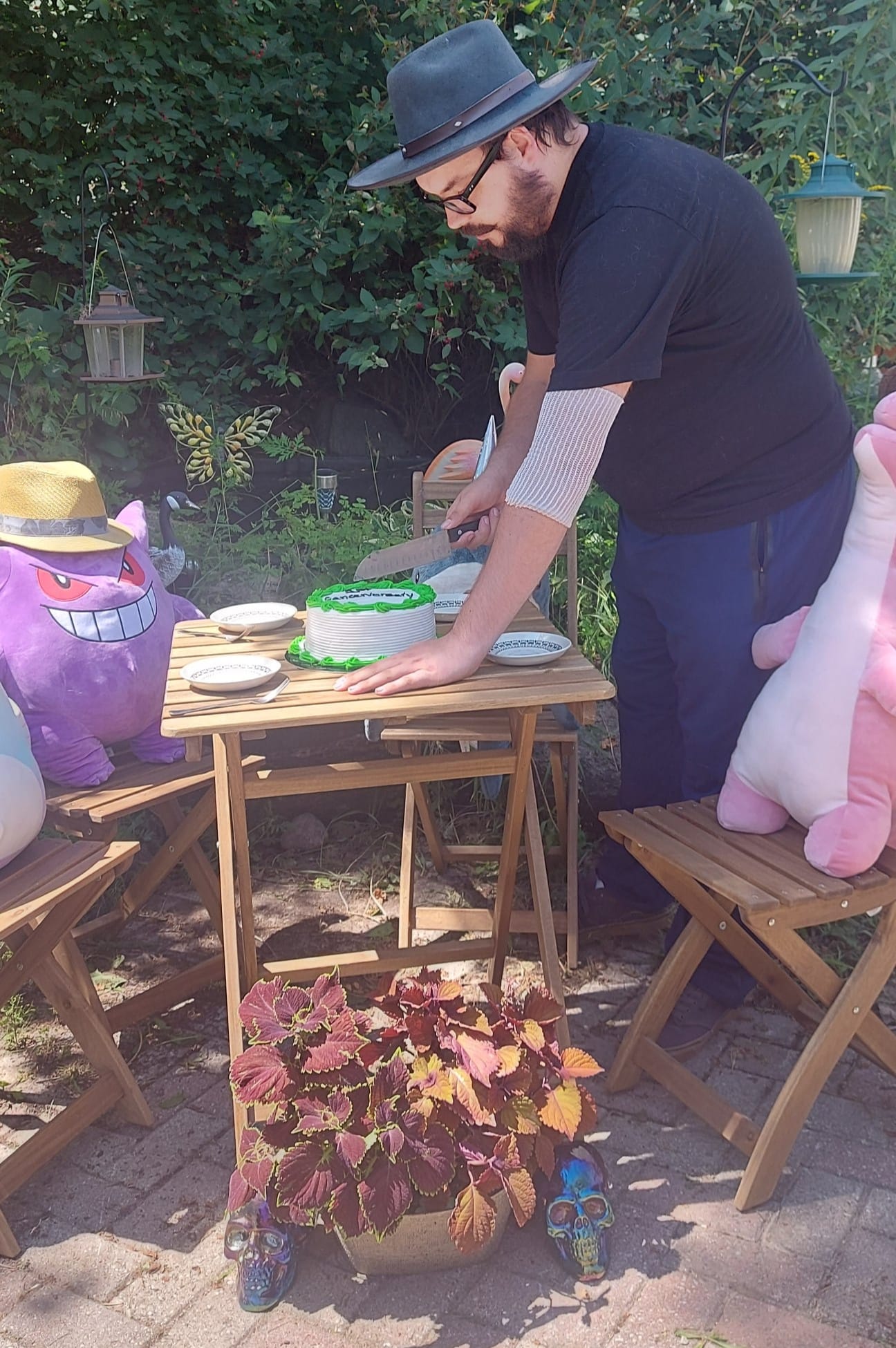 A man with facial hair and a felt sun hat has is cutting a white and green frosted cake at a stuffed animal patio party, Photo 1